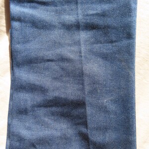 May include: Close-up of a section of blue denim jeans. The fabric shows subtle variations in color and texture, with a vertical seam running down the center. The denim has a classic, worn appearance.