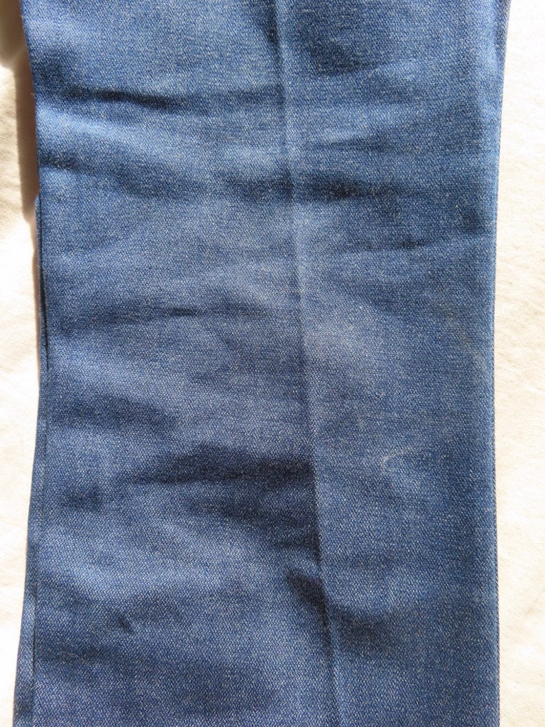 May include: Close-up of a section of blue denim jeans. The fabric shows subtle variations in color and texture, with a vertical seam running down the center. The denim has a classic, worn appearance.