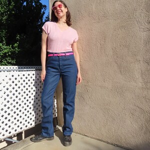 May include: A light pink V-neck top, dark blue jeans, and a bright pink belt. The jeans have a classic straight leg fit. The outfit is completed with black shoes and pink sunglasses.