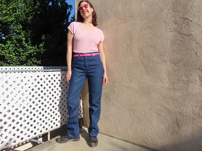 May include: A light pink V-neck top, dark blue jeans, and a bright pink belt. The jeans have a classic straight leg fit. The outfit is completed with black shoes and pink sunglasses.