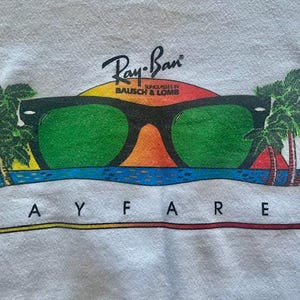 Pode incluir: Camisola branca com um gr&aacute;fico de &oacute;culos de sol Ray-Ban Wayfarer com lentes verdes, um fundo de p&ocirc;r do sol e palmeiras. O log&oacute;tipo Ray-Ban est&aacute; acima dos &oacute;culos.
