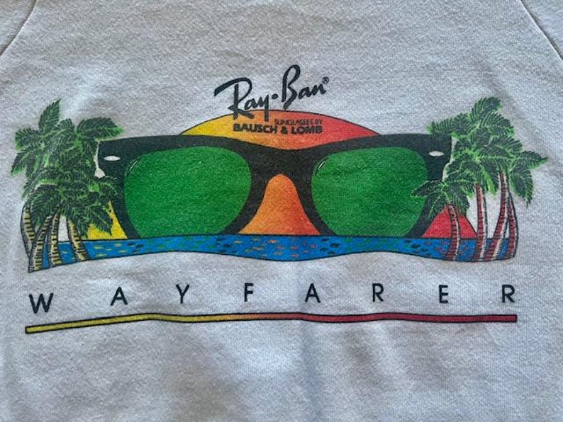Pode incluir: Camisola branca com um gr&aacute;fico de &oacute;culos de sol Ray-Ban Wayfarer com lentes verdes, um fundo de p&ocirc;r do sol e palmeiras. O log&oacute;tipo Ray-Ban est&aacute; acima dos &oacute;culos.