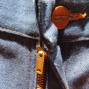 May include: Close-up of blue denim jeans with a copper-colored Wrangler button and a brass Scovill zipper. The stitching is in a contrasting orange-brown color, highlighting the seams and details of the jeans.