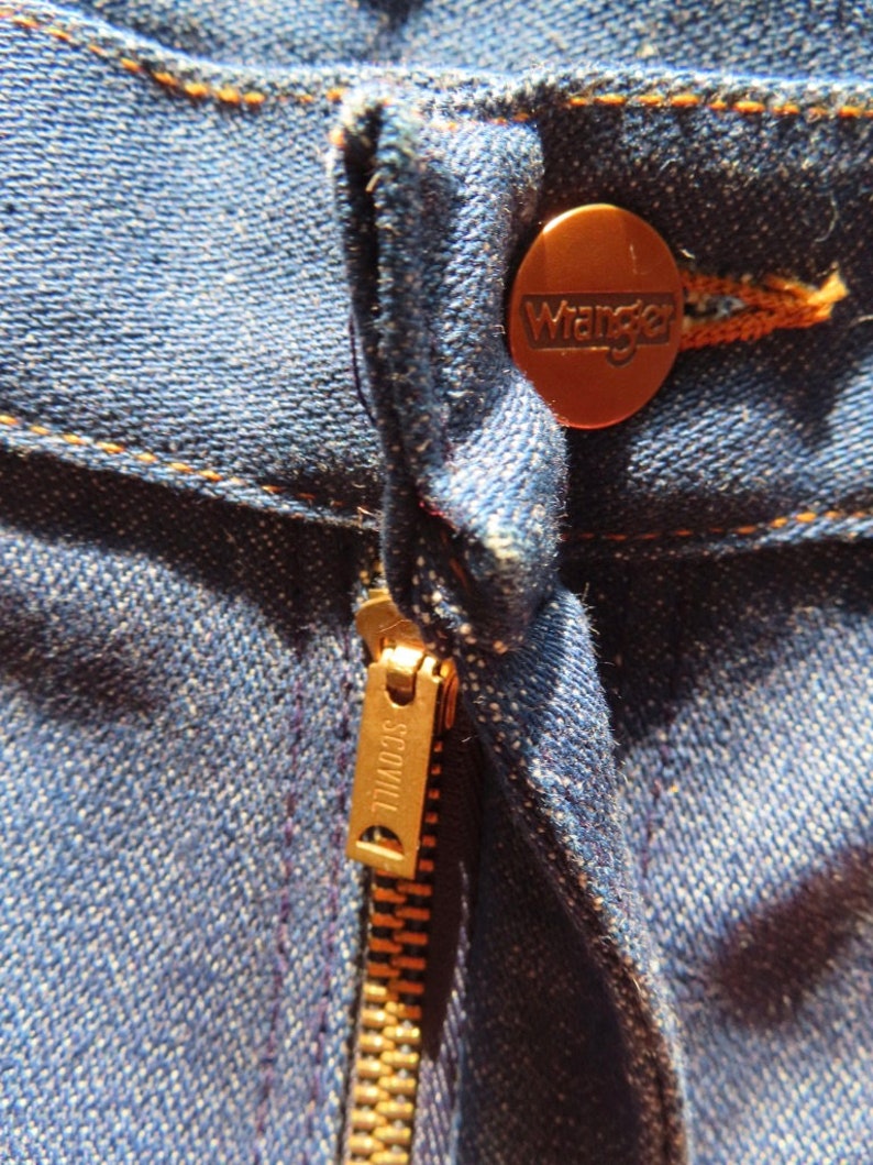 May include: Close-up of blue denim jeans with a copper-colored Wrangler button and a brass Scovill zipper. The stitching is in a contrasting orange-brown color, highlighting the seams and details of the jeans.