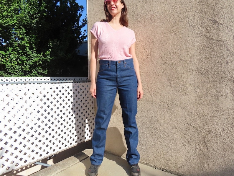 May include: A person wearing a light pink V-neck shirt and dark blue denim jeans. The jeans have a high waist and a straight leg. The person is wearing dark sneakers and pink sunglasses.