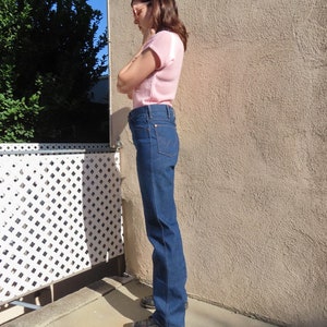 May include: Dark wash denim jeans with a classic straight leg fit. The jeans have a high waist and a traditional five-pocket design. The model is wearing a light pink short-sleeved top. The background includes a white lattice fence and a textured wall.