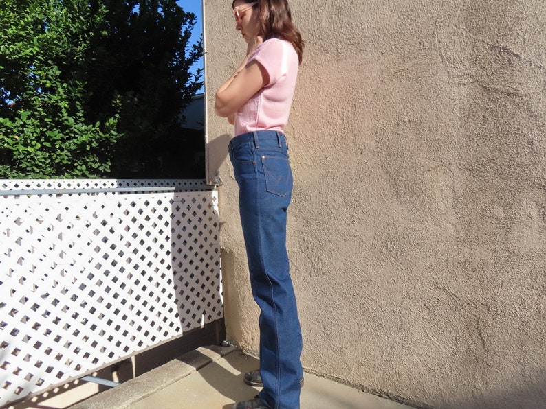 May include: Dark wash denim jeans with a classic straight leg fit. The jeans have a high waist and a traditional five-pocket design. The model is wearing a light pink short-sleeved top. The background includes a white lattice fence and a textured wall.