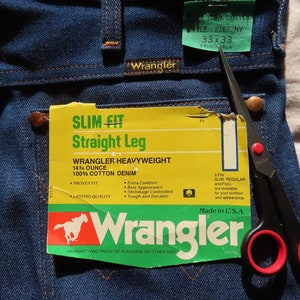 May include: Close-up of Wrangler blue denim jeans with tags. The tags read "SLIM FIT Straight Leg" and "WRANGLER HEAVYWEIGHT 14&frac34; OUNCE 100% COTTON DENIM". The jeans are size 33x33, with a waist of 33 inches and a 33-inch inseam.