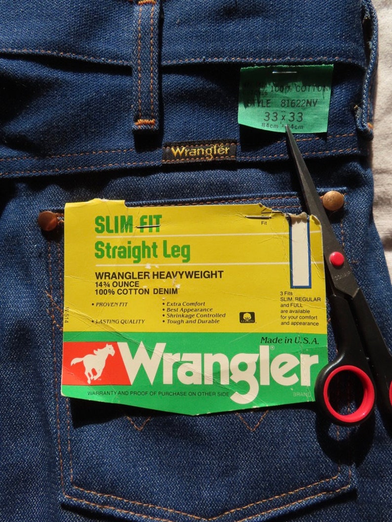 May include: Close-up of Wrangler blue denim jeans with tags. The tags read "SLIM FIT Straight Leg" and "WRANGLER HEAVYWEIGHT 14&frac34; OUNCE 100% COTTON DENIM". The jeans are size 33x33, with a waist of 33 inches and a 33-inch inseam.
