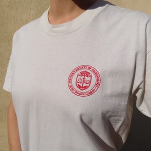 Peut inclure: Un t-shirt blanc avec un écusson rouge portant l'inscription "WESTERN SOCIETY OF CRIMINOLOGY" et "SOSU Student Chapter". L'écusson comprend une balance, un livre et un dessin de bâtiment. Le t-shirt a des manches courtes et un col rond.