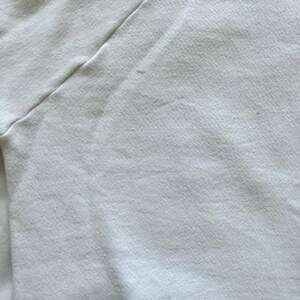 Pode incluir: Close-up de um tecido branco, possivelmente uma camiseta ou outra pe&ccedil;a de roupa. O tecido tem uma superf&iacute;cie ligeiramente texturizada com costuras vis&iacute;veis ao longo de uma costura diagonal. A cor &eacute; um branco sujo ou creme.