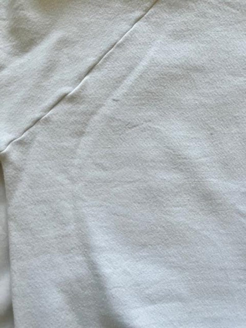 Pode incluir: Close-up de um tecido branco, possivelmente uma camiseta ou outra pe&ccedil;a de roupa. O tecido tem uma superf&iacute;cie ligeiramente texturizada com costuras vis&iacute;veis ao longo de uma costura diagonal. A cor &eacute; um branco sujo ou creme.