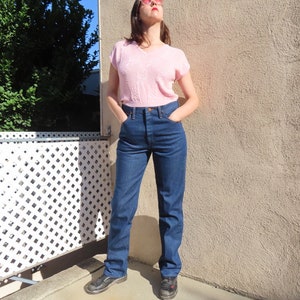 May include: A woman wearing a light pink V-neck short-sleeve shirt and high-waisted, dark blue denim jeans. The jeans have a classic straight-leg fit. The woman is wearing black shoes and sunglasses.