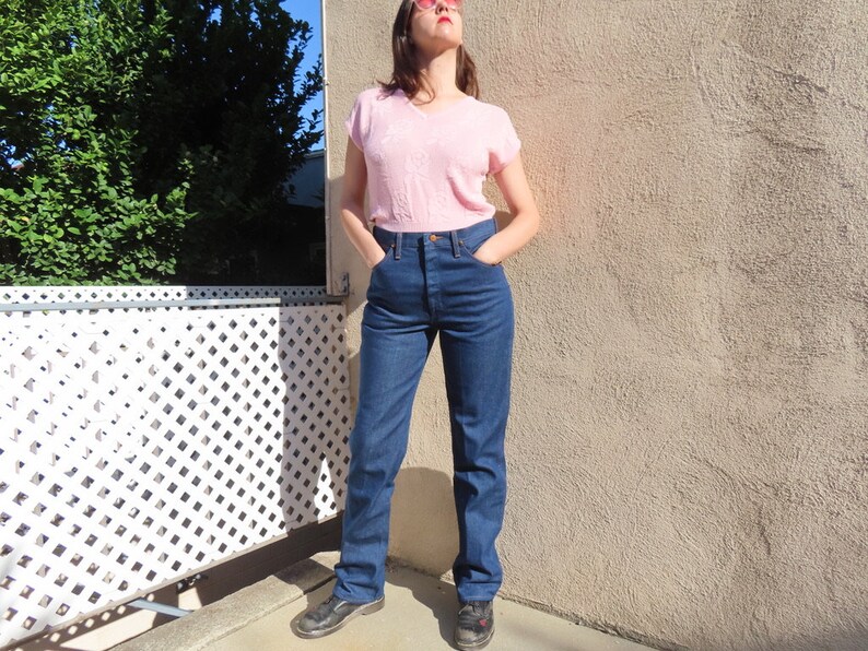 May include: A woman wearing a light pink V-neck short-sleeve shirt and high-waisted, dark blue denim jeans. The jeans have a classic straight-leg fit. The woman is wearing black shoes and sunglasses.