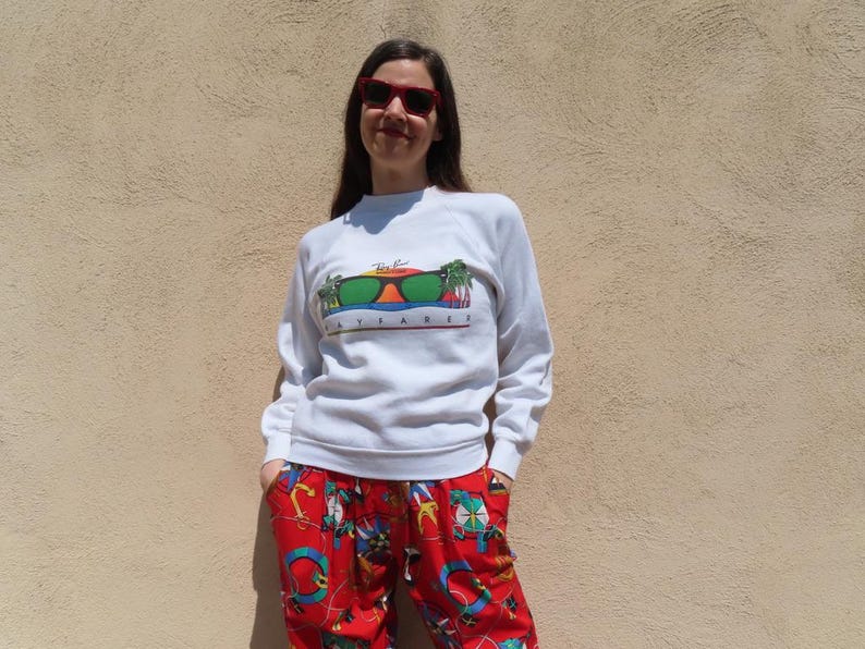 Pode incluir: Uma pessoa vestindo um moletom branco com um gr&aacute;fico de &oacute;culos de sol e o texto "Ray-Ban Apparel". A pessoa tamb&eacute;m est&aacute; usando &oacute;culos de sol vermelhos e cal&ccedil;as estampadas vermelhas.