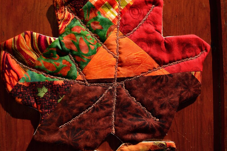 May include: A patchwork leaf-shaped quilt block with a variety of colorful fabrics, including red, orange, green, and brown. The fabric is sewn together in a geometric pattern and the edges are trimmed with gold thread.