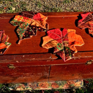 May include: Four quilted fabric leaf-shaped coasters in shades of red, orange, brown, and green, arranged on a wooden chest.