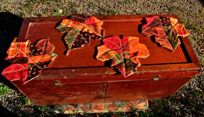 May include: Four quilted fabric leaf-shaped coasters in shades of red, orange, brown, and green, arranged on a wooden chest.
