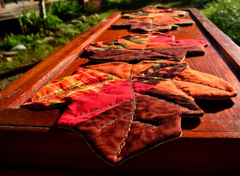 May include: A quilted table runner with a fall leaf design. The runner is made of various fabrics in shades of red, orange, brown, and green. The leaves are stitched together with a brown thread and have a beaded border.