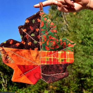 Könnte beinhalten: Ein bunter Patchwork-Topflappen in Form eines Ahornblattes, hergestellt aus verschiedenen Stoffen in Herbstfarben wie Orange, Rot, Braun und Grün. Das Blatt wird vor einem blauen Himmel und grünen Bäumen gehalten.
