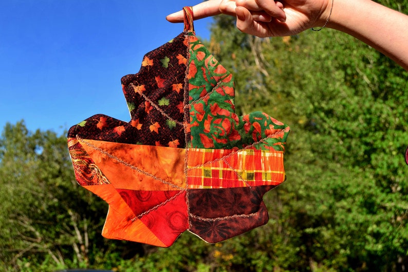 May include: A colorful patchwork maple leaf pot holder, made from different fabrics with autumnal colors like orange, red, brown, and green. The leaf is held up against a blue sky and green trees.