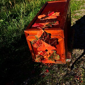 May include: A wooden storage chest with a fall-themed fabric garland of orange, red, and green leaves. The chest is sitting on a gravel surface.