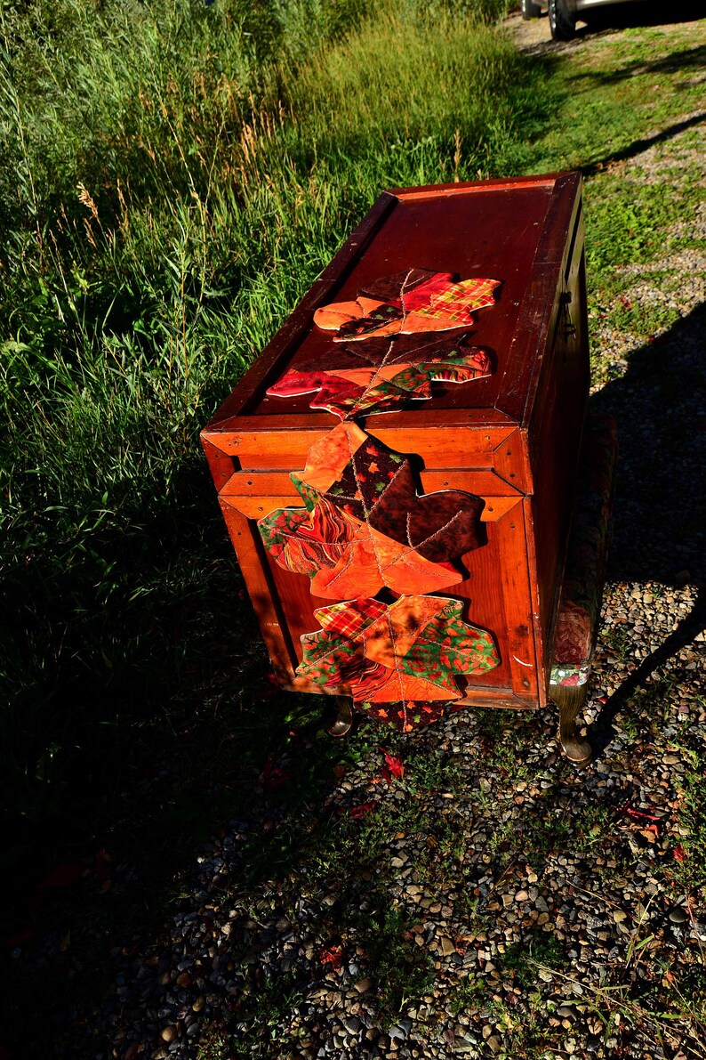 May include: A wooden storage chest with a fall-themed fabric garland of orange, red, and green leaves. The chest is sitting on a gravel surface.