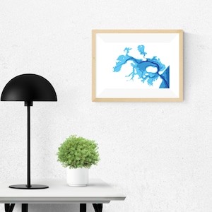 Pode incluir: Uma impressão artística emoldurada com um design de mapa abstrato azul e branco. A obra de arte é exibida em uma moldura de madeira clara e pendurada em uma parede branca. Uma lâmpada preta e uma pequena planta em vaso estão em uma mesa branca.