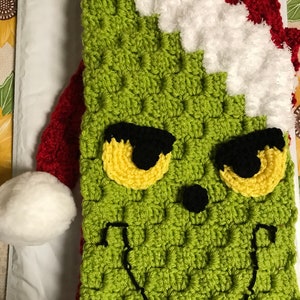 May include: A green crocheted Grinch-themed item with a red and white Santa hat design. The Grinch's face is detailed with yellow eyes, black pupils, and a stitched smile. The item is likely a holiday-themed accessory.