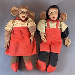 May include: Two vintage plush monkey dolls, one wearing a red dress and the other wearing red overalls. Both dolls have large ears and expressive faces.