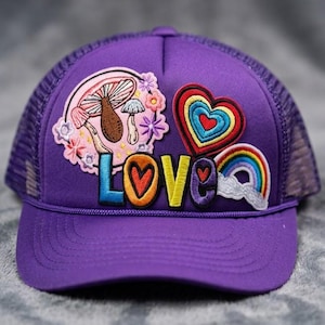 Puede incluir: Gorra de camionero morada con parches bordados. Los parches incluyen un diseño de hongo, un corazón, un arcoíris y la palabra "LOVE" en letras coloridas. La gorra tiene una parte trasera de malla y una visera curva.