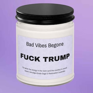 May include: A white candle in a glass jar with a black lid. The label reads "Bad Vibes Begone" and "FUCK TRUMP". The text below states the scent is Smudge-Grade Sage & Restorative Lavender.