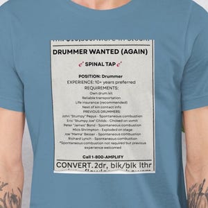 May include: A light blue t-shirt with a printed advertisement for a drummer position with Spinal Tap. The ad states "Drummer Wanted (Again)" and lists requirements and previous drummers. The shirt is worn by a person.