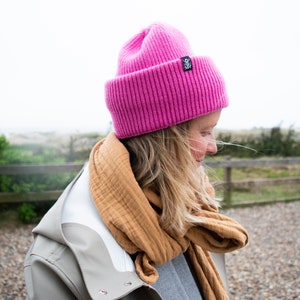 Könnte beinhalten: Eine Frau trägt eine rosa Mütze, einen braunen Schal und eine graue Jacke. Sie steht vor einem Holzzaun und einem Grasfeld.