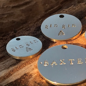 May include: Three round, gold-colored metal tags with engraved text. The first tag reads "gin gin" with a small tree design. The second tag reads "gin gin" with a small leaf design. The third tag reads ".BAXTER".