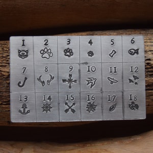 K&ouml;nnte beinhalten: Ein Stempelset aus Metall mit 18 verschiedenen Designs, nummeriert von 1 bis 18. Die Designs umfassen eine Katze, einen Pfotenabdruck, einen Knochen, einen Fisch, einen Haken, Geweih, einen Kompass, einen Vogel, ein Messer, einen Schmetterling, einen Anker, ein Steuerrad, Pfeile, eine Schneeflocke, ein Blatt und eine Beerenansammlung.
