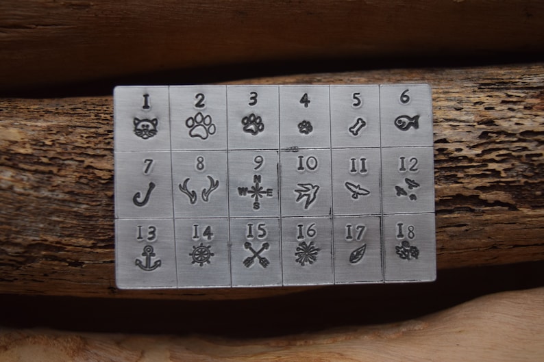 K&ouml;nnte beinhalten: Ein Stempelset aus Metall mit 18 verschiedenen Designs, nummeriert von 1 bis 18. Die Designs umfassen eine Katze, einen Pfotenabdruck, einen Knochen, einen Fisch, einen Haken, Geweih, einen Kompass, einen Vogel, ein Messer, einen Schmetterling, einen Anker, ein Steuerrad, Pfeile, eine Schneeflocke, ein Blatt und eine Beerenansammlung.