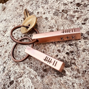 May include: Two rectangular copper keychains attached to a brass key ring. Each keychain features engraved tally marks, and one keychain includes the engraved text K + R. The metal surfaces have a warm, metallic copper tone with visible texture.