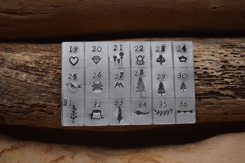 K&ouml;nnte beinhalten: Ein Stempelset aus Metall mit 36 verschiedenen Designs, darunter Herzen, Diamanten, Blumen, K&auml;fer, Kronen, V&ouml;gel, Totenk&ouml;pfe, Berge, B&auml;ume, Weizen und einen Baumstumpf. Die Zahlen 19 bis 36 sind &uuml;ber jedem Design aufgedruckt.
