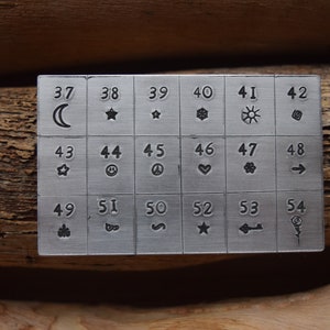 K&ouml;nnte beinhalten: Eine Metallplatte mit 18 nummerierten Quadraten, die jeweils ein anderes Symbol enthalten. Die Symbole umfassen einen Halbmond, einen Stern, eine Schneeflocke, eine Sonne, einen Wirbel, ein Smiley, ein Peace-Zeichen, ein Herz, eine Blume, einen Pfeil, einen Baum, ein Unendlichkeitszeichen, eine Schlange, einen Stern, einen Schl&uuml;ssel und eine Rose.