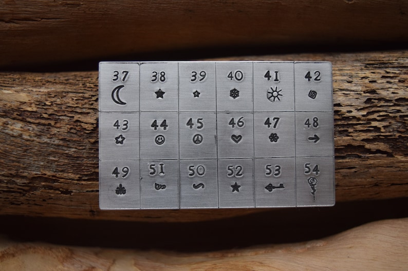 K&ouml;nnte beinhalten: Eine Metallplatte mit 18 nummerierten Quadraten, die jeweils ein anderes Symbol enthalten. Die Symbole umfassen einen Halbmond, einen Stern, eine Schneeflocke, eine Sonne, einen Wirbel, ein Smiley, ein Peace-Zeichen, ein Herz, eine Blume, einen Pfeil, einen Baum, ein Unendlichkeitszeichen, eine Schlange, einen Stern, einen Schl&uuml;ssel und eine Rose.