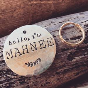 K&ouml;nnte beinhalten: Ein runder, messingfarbener Hundemarkenanh&auml;nger mit dem Text "hello, i'm MAHNEE" und einem kleinen Blattdesign. Der Anh&auml;nger ist an einem goldenen Metallring befestigt.