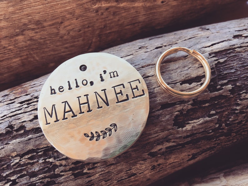 K&ouml;nnte beinhalten: Ein runder, messingfarbener Hundemarkenanh&auml;nger mit dem Text "hello, i'm MAHNEE" und einem kleinen Blattdesign. Der Anh&auml;nger ist an einem goldenen Metallring befestigt.