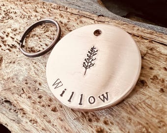 May include: A round, copper-colored metal tag featuring a stamped image of a willow branch and the name Willow in capital letters. The tag includes a small hole at the top and is attached to a circular metal split ring for key attachment.