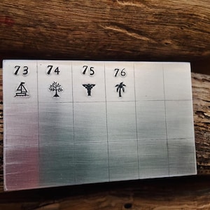 K&ouml;nnte beinhalten: Eine Metallplatte mit f&uuml;nf nummerierten Quadraten, die jeweils ein anderes Symbol enthalten: ein Segelboot, einen Baum, ein medizinisches Symbol, eine Palme und die Nummer 76.