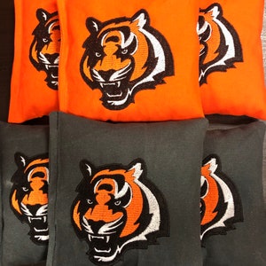 May include: Six bean bags with a tiger logo embroidered on each. The bean bags are made of two different fabrics, one orange and one gray. The tiger logo is black and white with orange accents.