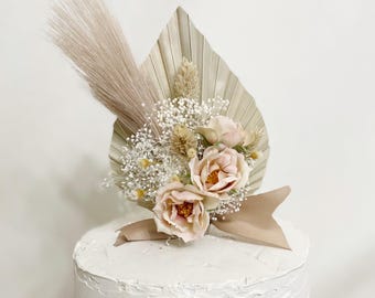 May include: A cake topper featuring a beige palm leaf, dried flowers, and blush-colored roses. A beige ribbon is tied around the flowers. The cake is white with a textured surface. The website address TheVirginiaArtisans.com is visible.
