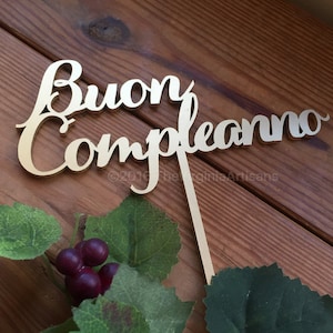 May include: A gold-colored cake topper with the Italian phrase "Buon Compleanno" in a cursive font. The topper is on a wooden surface with green leaves and purple berries.