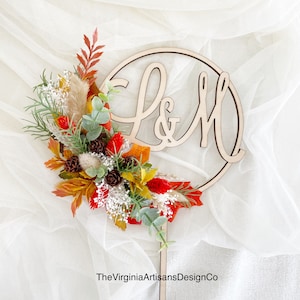May include: A wooden wedding cake topper featuring the letters "L & M" within a circle, adorned with a colorful arrangement of fall-themed flowers, leaves, and pinecones. The topper is on a wooden stick.