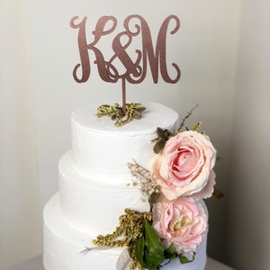 May include: A three-tiered white cake decorated with two large pale pink roses and green foliage. A copper-colored cake topper featuring the letters K&M in a cursive script is inserted into the top tier of the cake, centered above the flowers.